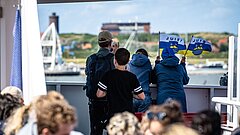 Anreise nach Juist mit der Inselfähre in Richtung Hafen, Kinder schauen auf dem Oberdeck der Inselfähre in Richtung Hafen, in den Hänfen Juist Fahnen, im Vordergrund unscharfe Menschen
