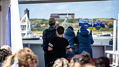 Anreise nach Juist mit der Inselfähre in Richtung Hafen, Kinder schauen auf dem Oberdeck der Inselfähre in Richtung Hafen, in den Hänfen Juist Fahnen, im Vordergrund unscharfe Menschen