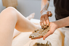 Thalassoangebote auf Juist - Foto zeigt beide Hände einer Therapeutin, die Schlick verribt, im Hintergrund das Bein einer Patientin auf einer Wellnessliege