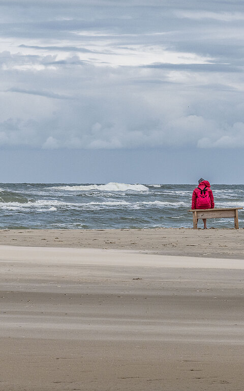 Alleinreisende und Singles auf Juist im Urlaub - Singleurlaub auf Juist - Bild zeigt eine Person in einer roten REgenjacke auf einer Badnk direkt an der Wasserkante am Strand auf Juist