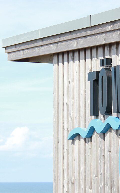 Aussenansicht TöwerVital auf Juist mit schwarz-blauem Schriftzug auf hölzerner Fassade, Strand mit Meer unscharf im Hintergrund