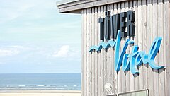 Aussenansicht TöwerVital auf Juist mit schwarz-blauem Schriftzug auf hölzerner Fassade, Strand mit Meer unscharf im Hintergrund