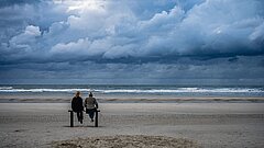 Wasserkante der Nordsee auf Juist am Strand mit blick auf zwei Personen auf einer Bank und bewölktem Himmel